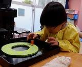 幼児が絵の具がついたピーマンを画用紙の上に乗せ、ピーマンを見つめている写真