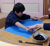幼児が両手を広げて書き初めをする画用紙の大きさを確かめている写真。