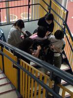 口をハンカチで押さえながら車椅子に座った生徒を、4名の教員が担いで、体育館の階段を下りて避難している写真。