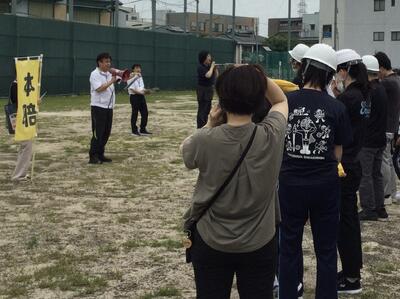 避難集合場所の運動場に集まった児童生徒と教員が、校長先生の話を聞いている写真。